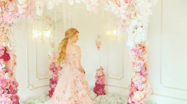 Fantezi mutlu prenses beyaz odada çiçek açan bahar manzarası, çiçek desenli kemerler yükseldi. Kadın kraliçe sarı saçlı, klasik pembe gece elbisesi arkadan görünüşlü. Neşeli bayan geriye bakıp gülümsüyor. 4k