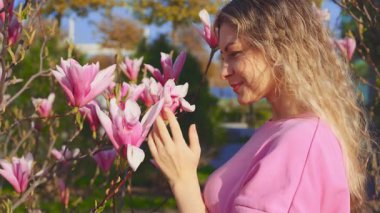 Portre kıvırcık sarı saçlar mutlu kadın eli pembe manolya çiçeği tomurcuğuna dokunuyor doğal güzellik tatlı kız gülümseyen yüz kokusu bahar bahçesinin tadını çıkarıyor yeşil ağaç güneş ışığı parkı 4k.
