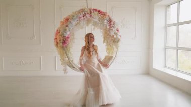 Mutlu fantezi kadın sarışın çiçek salıncağında oturmuş aşkı bekliyor. Romantik prenses mankeni seksi uzun elbise. Sarı saçlar. Beyaz klasik oda çiçekli dekor asılı sandalye 4k