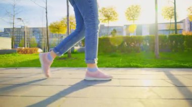 Yakın plan kadın bacakları modern şehrin kaldırım caddesinde yürüyor. Kamu parkında yürüyen bir kadın, gün batımında yeşil çimen yaz ağaçlarının dışında bahar ışığı. Pembe spor ayakkabılı, kot pantolonlu kız.