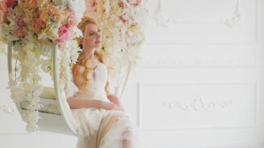 Mutlu gelin sarışın, pembe çiçekli taht koltuğu salıncağında oturuyor. Seksi manken beyaz gelinlik. Uzun sarı saçlı, zarif, bol saçlı, saç stili, klasik boho tarzı.