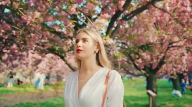happy woman blonde long hair walking in park sakura pink flowers branch tree enjoy fantastic spring nature day cherry blossom sun light. adult girl fashion model beauty smiling face, white dress 4k