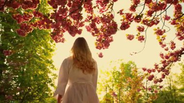 Happy young woman spinning dancing enjoy spring nature green leaves trees pink sakura flowers sun light park. Beauty cheerful girl smiling face, long blond hair flying in wind motion. White dress 4k