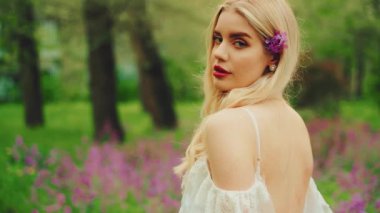 Art portrait beauty face girl blue eyes, gentle makeup red full lips. young woman enjoy summer green nature. white long dress boho style open bare back. Blonde hair with purple flowers sexy lady. 4k