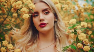 Woman beauty face close-up eyes lips in yellow flowers. sexy girl fashion model looking camera. spring garden tree enjoy smell. Blonde curly hair lady inhales floral scent white dress boho style. 4k