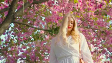 fantasy young happy woman blonde hair red lips beauty smiling face walking at public park sakura pink flowers. Adult girl sexy fashion model. spring garden tree nature day sun light. art 4k 8 march 