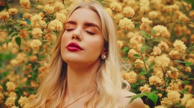 Fantasy sexy girl fairy in spring garden flowering tree. happy woman enjoy smell yellow flowers. white dress. Blonde curly hair lady inhales floral scent. Beauty face natural makeup pearl earrings. 4k