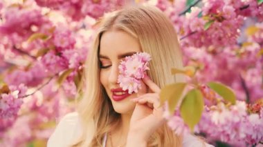 Portrait young happy woman blonde hair beauty smiling face look at camera blue eyes holds sakura pink flowers in hand. Adult girl fashion model perfect skin natural makeup. spring tree nature day eco