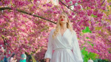 young happy woman blonde hair beauty smiling face walking at public park enjoying sakura pink flowers. Adult girl sexy fashion model. spring garden tree nature day sun light. art 4k. 8 march holiday