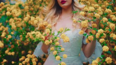 Fantasy sexy girl in spring garden flowering tree. happy woman enjoy smell yellow flowers. white dress. Blonde curly hair lady in floral scent. Beauty face cropped natural makeup red lips close-up. 4k