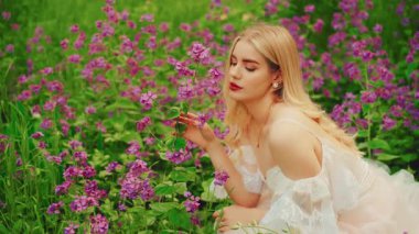 beauty face girl fairy sitting in summer fied flowering meadow green grass. happy woman forest nymph hand touching purple flowers. white dress boho style. Blonde hair sexy lady inhales floral scent.
