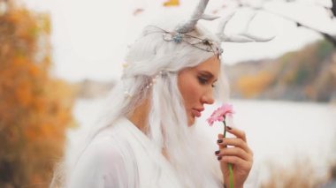 Fantezi genç elf kadın pembe Gerbera çiçeği tutuyor. Güzel yüzlü, seksi gözlü, peri dudaklı, boynuzlu, sarı saçlı, küllü, beyaz elbiseli. Efsanevi peri kadın çiçek kokusu soluyor..