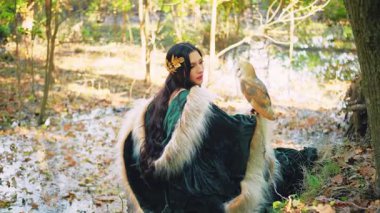 Beyaz kuşlu orman perisi fantezi kadın, baykuşu elinde tutuyor yere düşmüş yeşil yaprakların üzerinde oturuyor, ortaçağ tarzı. Orman sonbahar ağaçları doğa nehir ışığı. Uzun siyah saçlı, altın sarısı.