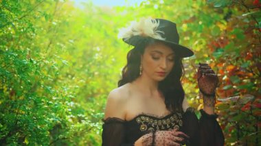 Kameraya bakan mutlu kadın portresi, eski moda siyah şapkalı asil bayan seksi kız Victoria tarzı eski moda eski moda elbise peçe eldivenleri. Yeşil ağaçlar orman güneşi