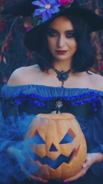 Cadılar Bayramı portresi mutlu gülümseyen yüzlü cadı kadın. Mor kostümlü kız siyah külah şapkalı. Elinde turuncu Jacko fener kabağı tutan gotik bayan mavi dumanlı sonbahar arka plan dikey video