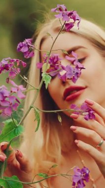 Güzel, gülümseyen kız peri masalı meleği. Mutlu kadın yaz yeşili tarla işçisinin mor çiçeklere dokunuşunun tadını çıkarıyor. Boho tarzı beyaz elbise. Sarışın, seksi bayan çiçek kokusu soluyor. dikey video