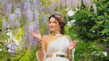 Gelin kadın bahar bahçesinde yürüyor mor salkım çiçeklerine dokunuyor, yeşil ağaç yaz doğasına. Seksi kız kraliçe antik Yunan tarzı beyaz ipek elbise, altın sarısı diyabet. Fantezi tanrıçası gülen surat..