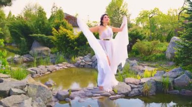 Fantezili mutlu kadın, taşın üzerinde, nehir elleriyle gökyüzüne poz veriyor. Beyaz ipek uzun elbise. Eski tarz rüzgârda uçuşuyor. Seksi lüks gelin kız Yunan tarzı kıyafetler. Su Gölü nehri güneşi yaz günü 4k