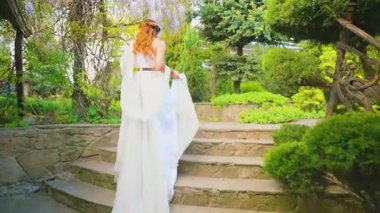 Fantezi kadın Yunan tanrıçası bahar bahçesinde yürüyor mor çiçekler yeşil ağaç doğa güneş ışığı. Seksi kız, antika tarzı beyaz ipek elbise, kızıl saçlı altın sarısı, bayan gelinin arkadan görünüşü 4k.