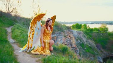 Fantezi kadın melek diz çöküp dua eder ve yükselir, altın beyaz kanatlar uzun altın elbiseler dağ manzarasının tepesinde durur kutsal mavi gökyüzü günbatımında. Sanat perisi kız seksi model 4k