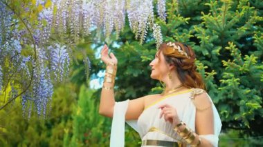 Fantezi kadın Yunan tanrıçası bahar bahçesinde yürüyor, eli mor salkımcık çiçeklerine dokunuyor, yeşil ağaç yaz doğası güneş ışığına. Seksi kız, antika tarzı beyaz ipek elbise, altın sarısı 4k mücevher.