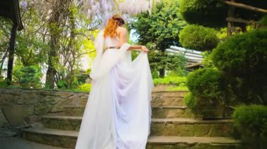 Fantezi kadın Yunan tanrıçası bahar bahçesinde yürüyor mor çiçekler yeşil ağaç doğa güneş ışığı. Seksi kız, antika tarzı beyaz ipek elbise, kızıl saçlı altın sarısı, bayan gelinin arkadan görünüşü 4k.