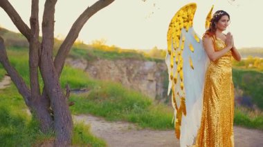 Fantezi mutlu kadın gözleri kapalı başmelek dua ederken ellerini kavuşturuyor. Beyaz, altın kanatlı, melek kostümlü, metal, altın mücevherli taç takan. Seksi kız melek stili. Yaz doğası ilahi sihirli güneş ışığı gökyüzü