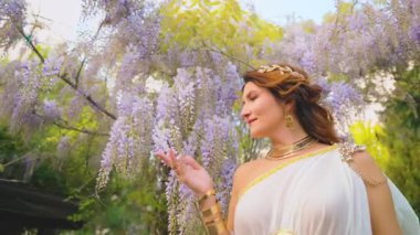 Fantezi kadın Yunan tanrıçası bahar bahçesinde yürüyor, eli mor salkımcık çiçeklerine dokunuyor, yeşil ağaç yaz doğası güneş ışığına. Seksi kız, antika tarzı beyaz ipek elbise, altın sarısı 4k mücevher.