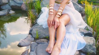 Fantezi kadın tanrıça nehir perisi yakın plan eller tene dokunur ellerle pürüzsüz bacaklar. Altın bilezikler, metal kolye, Yunan tarzı takılar. Kraliçe kız model beyaz elbise 4K