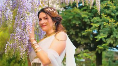 Gelin kadın bahar bahçesinde yürüyor mor salkım çiçeklerine dokunuyor, yeşil ağaç yaz doğasına. Seksi kız kraliçe antik Yunan tarzı beyaz ipek elbise, altın sarısı diyabet. Fantezi tanrıçası gülen surat.