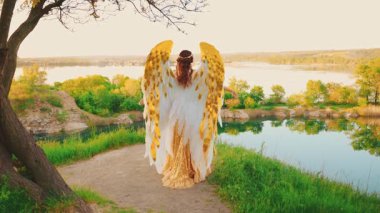 Silüet fantezi kadın tanrıça melek, dağın tepesinde duran altın beyaz kanatlı, altın sarısı elbisesiyle ellerini kaldırıyor ve nehrin sihirli mavi gökyüzüne geri dönüp dua ediyor. Sanat Perisi Kız Seksi Kadın