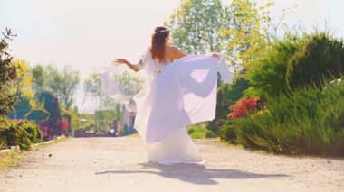 Mutlu kadın Yunan tarzı kıyafet arka bahçedeki patikada yürüyor seksi stil sahibi bayan arkadan bakıyor. Uzun beyaz elbiseli kadın. İpek tren kumaşı. Uçan rüzgar. Kız modasına uygun gelin. Gündüz güneşi yavaş çekim