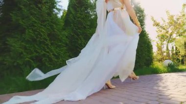 Yakın plan çekimde kadın bacakları yüksek topuklu seksi ayaklar giyerek patika boyunca yürüyor. Yunan kadın tanrıçası uzun beyaz elbiseli ipek trenle eski usul uçuyor. Kız modasına uygun gelin. Gün ışığı geri ışık