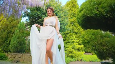 Gelin kadın bahar bahçesinde yürüyor mor salkımcık çiçekleri, yeşil ağaç yaz doğası güneş ışığı. Seksi kız kraliçe antik Yunan tarzı beyaz ipek elbise, altın sarısı diyabet. Fantezi kadın merdivenlerden 4K aşağı iniyor.