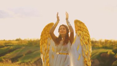 Fantezi kadın melek, dua eden güzel beyaz kanatlarıyla ellerini havaya kaldırıyor. Saha sihirli güneş parlak gökyüzü yeşil çim yar tepeleri. Uzun elbiseli melek kız kızıl saçlı. Peri kraliçesi seksi bayan. 4k