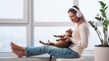 Mutlu sarışın kadın pencere kenarına oturur, panoramik kulaklıklar takar, kedi gibi dans eden kedilerle neşeli sürüş müziği dinler. Neşeli kız model, komik disko hayvanıyla dans etmekten hoşlanıyor..