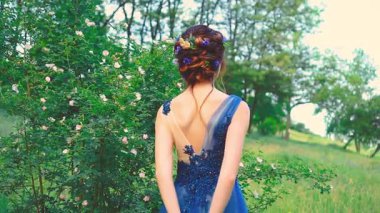 Fantezi kadın kraliçesi dikiz aynasından zarif, klasik saç stilini çiçeklerle topladı. Kız prenses doğanın tadını çıkarıyor. Yeşil yapraklı ağaç tarlası. Mavi romantik gece elbisesi bayan model