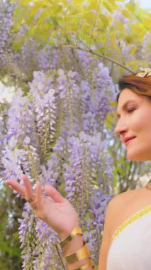 Fantezi kadın Yunan tanrıçası bahar bahçesinde yürüyor, eli mor salkımcık çiçeklerine dokunuyor, yeşil ağaç yaz doğası güneş ışığına. Seksi kız, antika tarzı, beyaz elbise, altın sarısı diyabet. dikey video.