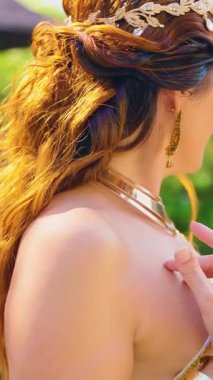 Fantezi kadın Yunan tanrıçası bahar bahçesinde yeşil ağaçta oturuyor yaz güneşi ışığı. Seksi kız kraliçe tacı beyaz elbise. Kızıl saçlar kıvırcıklaşır. Eski saç arkadan görünür. resim dikey video.