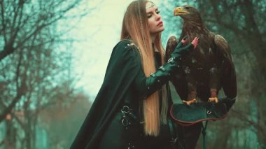 Hayvanlarla yaratıcı çekim süreci, kuşlarla moda modası. Beyaz kuyruklu kartal eli tutan kadın. Elf savaşçısı güzel yüz kahverengi kuş evcil hayvan kraliçesi siyah kostüm siyah ağaçlar 4k