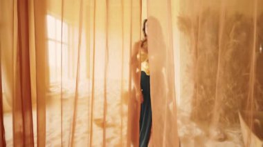 fantezi seksi kadın kum dolu bir odada yürüyor. Sarışın, güzel surat. Siyah uzun elbiseli manken kız altın korse eski tarz. Kadın eli açılır, perdeler ipekten kumaş atar. resim videosu 4k