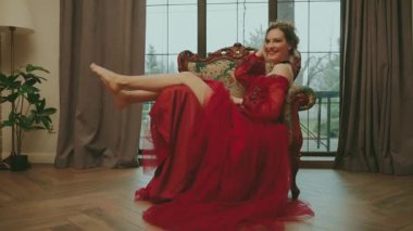 Sanat portresi mutlu, fantezi kızı prenses eski tarz tahta uzanmış çıplak bacakları sallıyor, neşeli bir ruh hali içinde. Sandalyede oturan genç seksi sarışın kadın. Klasik oda kırmızı kraliyet giysisi taç..