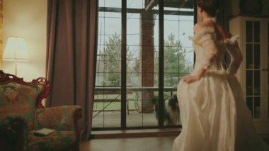 Genç, seksi sarışın kadın klasik odada koşuyor, pencereye gidip aşkı bekliyor. Peri masalı prenses gelin yetişkin kız arkadan güzel görünüm. Kraliyet evi beyaz eski moda elbise uzun balo elbisesi eski tarz 4K