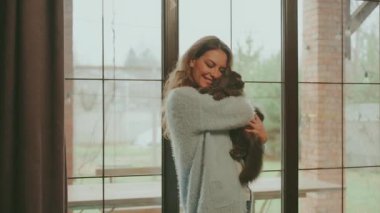 Mutlu kadın, sevimli, tüylü, gri kedi şefkatinin komik duygularına sarılıyor. Yetişkin, sarışın, güzel, gülümseyen bir kız. Rahat bir odada, rahat giysiler içinde. Ağızlık takan bir kadın.