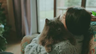 Mutlu kadın, sevimli, tüylü, gri kedi şefkatinin komik duygularına sarılıyor. Evde gülümseyen yetişkin güzel kız rahat bir odada örgü örüyor, ağızlık takıyor, el ele tutuşmuş hayvan öpüyor pencere kenarına oturuyor..