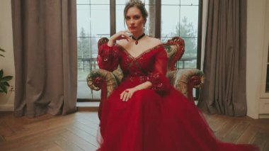 Fantezi kadın ortaçağ kraliçesi eski tarz eski moda sandalyede oturan, ortaçağ kızıl elbiseli kadın Gotik taç. Kraliyet klasik beyaz oda penceresi. Seksi Prenses kız soylu peri masalı Lady 4k.