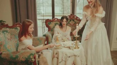 Gülümseyen mutlu kadın çaydanlığı tutup altın renkli bardak setine sıcak çay döker. Kahve masasındaki kanepede oturan asil kadınlar çay içerler. Oturma odasındaki dinlenen bayan beyaz, eski tarz elbise..