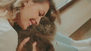 Camın kenarında oturan, elinde tüylü gri bir kedi tutan sevimli bir kadın. Neşeli kız duyguları. Aşk bakımı, gülümseyen hayvan ağızlık kürkü. Konforlu ev kıyafetleri. 4k