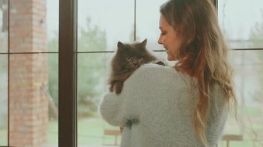 Mutlu sarışın kadın pencerenin kenarında durmuş elinde güzel tüylü gri kediyi tutuyor, neşeli kız sevgiyle bakıyor, gülen surat hayvan ağızlık kürkünü öpüyor. Konforlu ev kıyafetleri