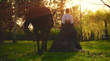 Atlı mutlu kadın fantezisi. Model kız dikiz aygırıyla yürüyor. Peri Kraliçe 'nin uzun etekli elbisesi rüzgarda uçuşuyor. Günbatımı ormanı yeşil ağaçlar sihirli güneş ışığı 4k